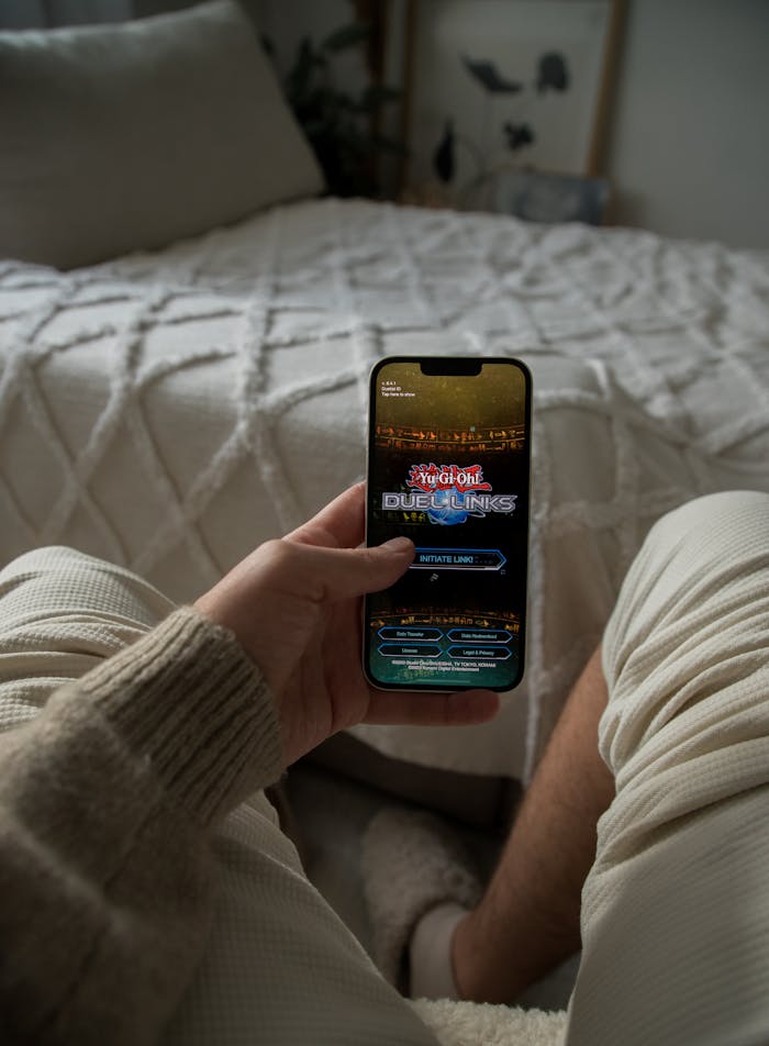 Hand holding smartphone in a cozy bedroom, displaying a gaming app.