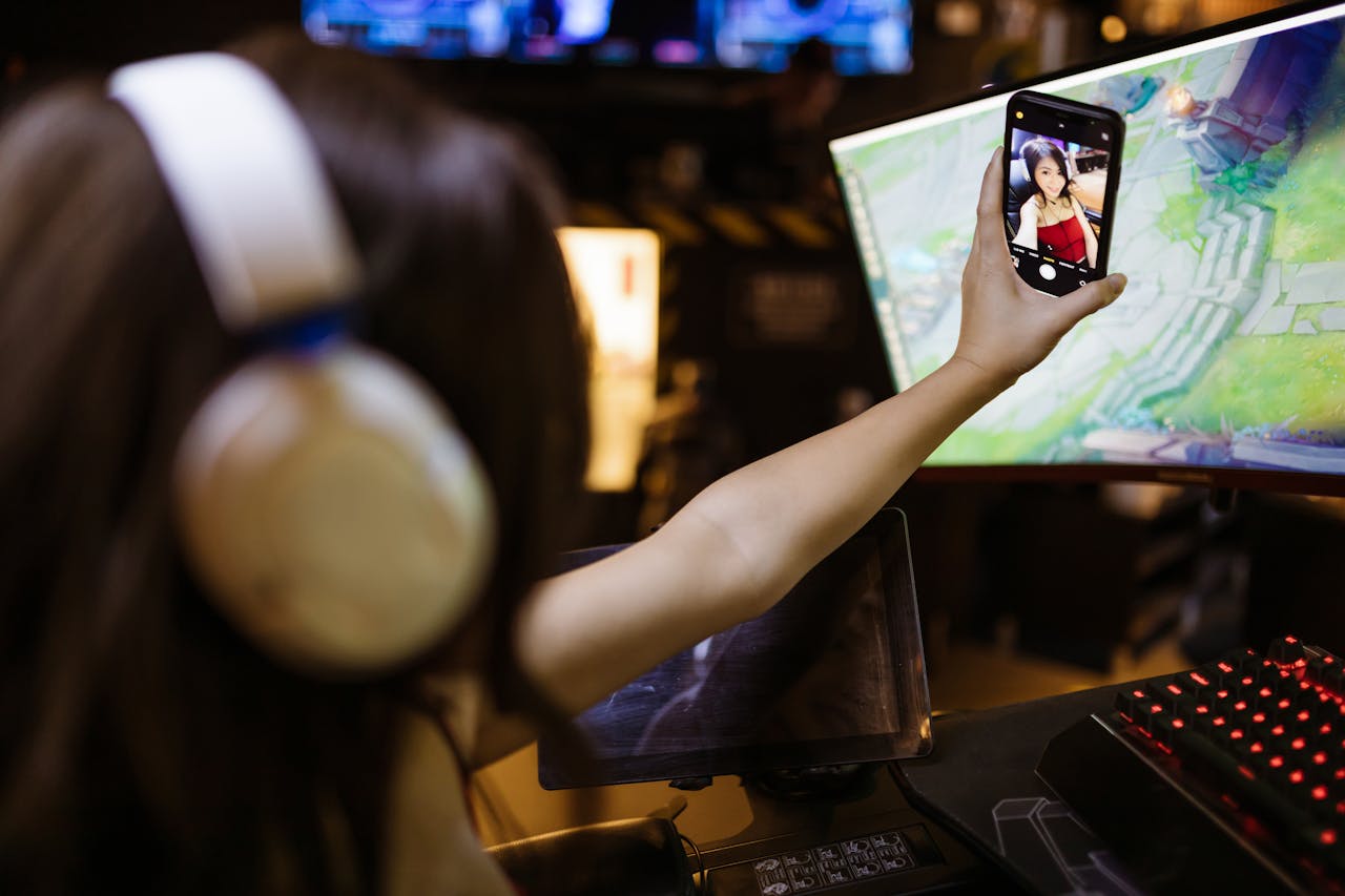 A woman wearing headphones takes a selfie with her smartphone while engaged in gaming indoors.